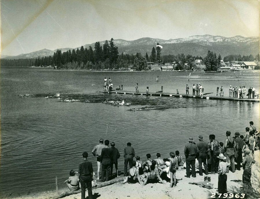 A swimming race occuring in McCall, Idaho where three CCC Camps converged to hold a carnical. A line of swimmers are in the water while spectators line both the shore and the peir where the race began. The town of McCall can be seen across the lake. Description reads: 'Swimming race CCC water Carnival at McCall, Idaho. K.D. Swan - 1933'.