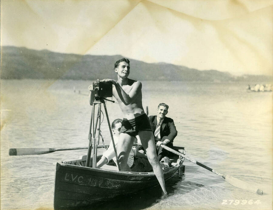 Three CCC men in a boat on the shore of a lake. One of the men is in a suit and tie and holds both oars. Another man, in swimming trunks, is standing half in the shallows and half in the boat. He is holding a camera that is standing on a tripod on the boat. The lettering on the side of the boat reads: 'L.V.C. N3'. Description reads: 'New York takes a hand at the camera. K.D. Swan - 1933.'