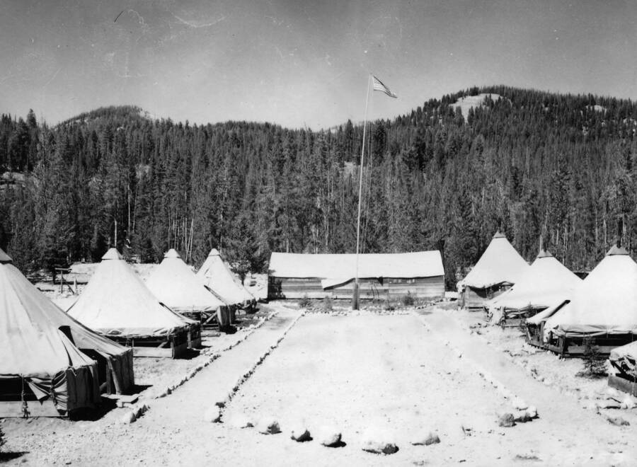 Two rows of tents and a headquarters wooden building with a flagpole in front of it. A clearing between the two rows of tents is bordered by rocks. A wooded hill rises in the background.