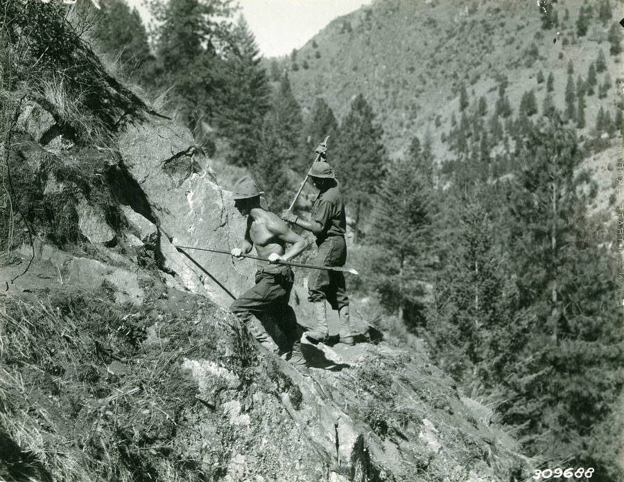Two CCC men working on the rocky slope of a canyon. Both are using hand tools. Description reads: 'Swan - Payette. CCC boys cleaning holes preparatory to loading Salmon river road job. July, 1935.'