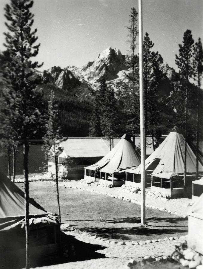 A mountain rises in the background behind a CCC Camp. The camp is made up of several tents and a wooden building behind a flagpole. The clearing in the center is bordered by rocks.