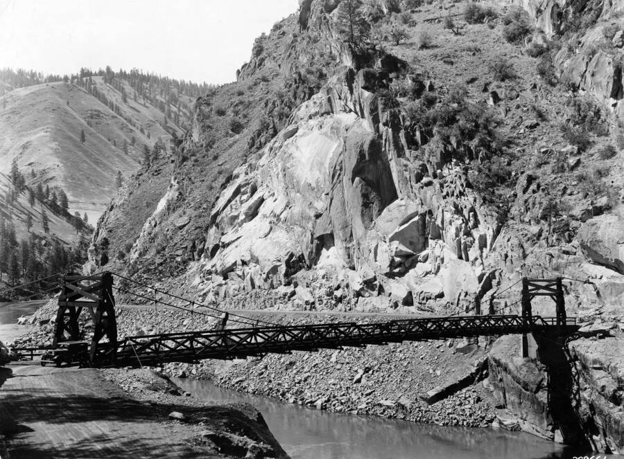 Manning Bridge spanning the Salmon Riverwith a cliff face and hill rising in the background. Description reads: 'Manning Bridge across Salmon River, built by CCC boys in 1934. Forest: Payette, State: Idaho, Date: 7/1935, Author: K.D. Swan'.