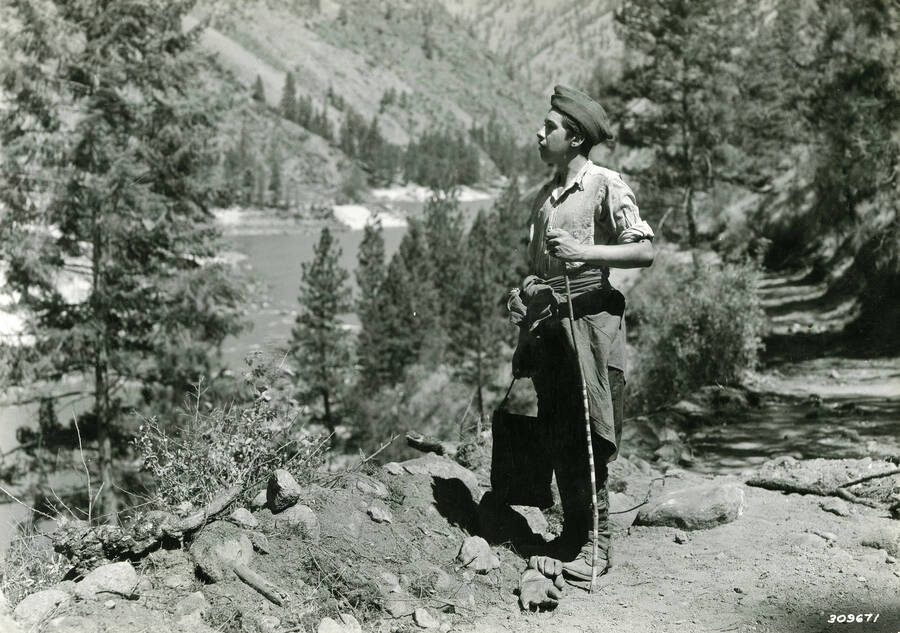 CCC man standing on a dirt path looking out across a river valley while leaning on a walking stick. Writing above the photo reads: 'Payette N.F., by K.D. Swan, CCC Waterboy on Salmon River road job. July 1935'.