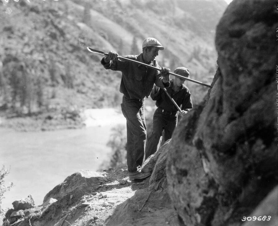 Two CCC Men working on the Salmon River road project using hand tools on the cliff face above the river.