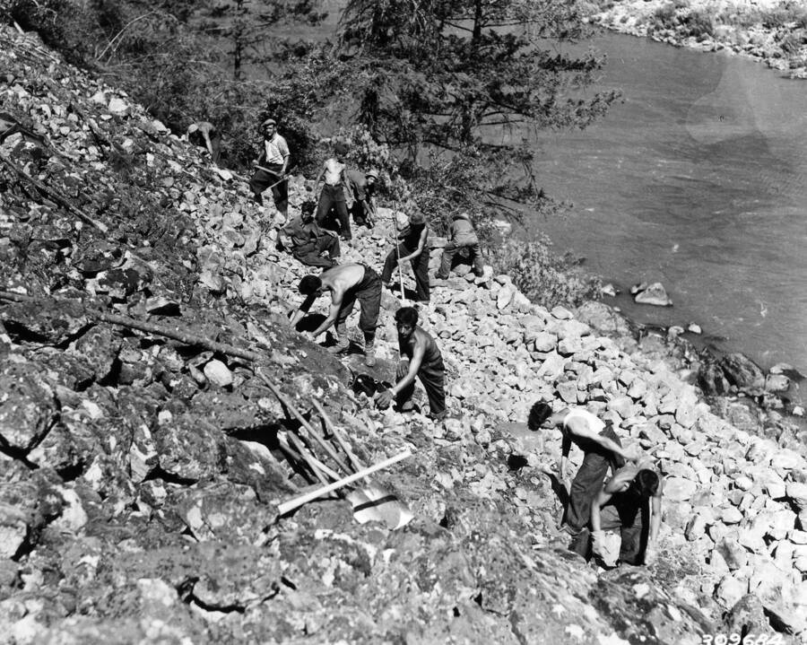 A CCC work crew working on a rocky hillside. Above them is a small pile of shovels.