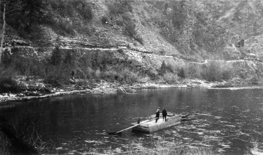 Two men standing on a boat on the river. Snow dusts the bank and hill on the other side of the river and a line runs along the hill that is presumably a road.