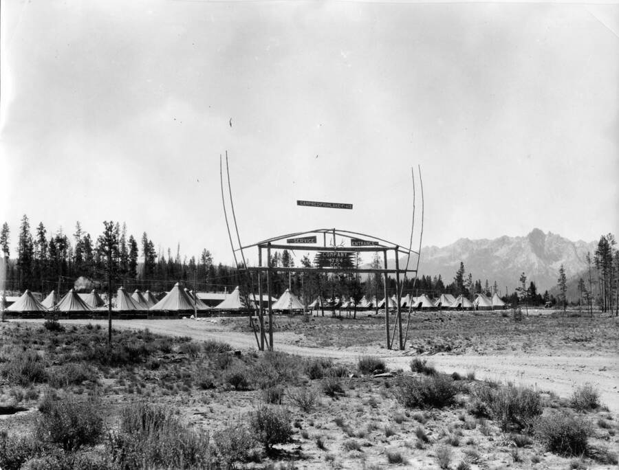 A CCC Camp sign stands in the foreground with a CCC tent camp in the background. The sign reads: 'Camp Red Fish Lake F-412 Service Entrance Company 4784 CCC'. The camp is surrounded by scrubland and woods with a mountain rising in the background.