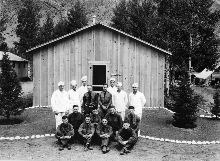 A group of CCC men pose for a group photo outside of a building labeled 'mess hall'. Some are in uniform, others are dressed as cooks. There is some nice landscaping outside the building, including two pine trees and a border of white rocks.