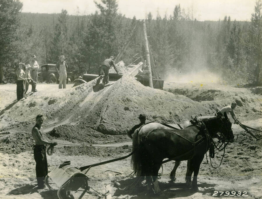 CCC Men and a team of horses loading gravel into trucks. The horses are resting in the foreground, while one man pushes a wheelbarrow full of gravel up a ramp while several other men look on. Description reads: 'CCC's load gravel for road to Redfish Lake. Forest: Sawtooth N.F., State: Idaho, Date: 1930's'.