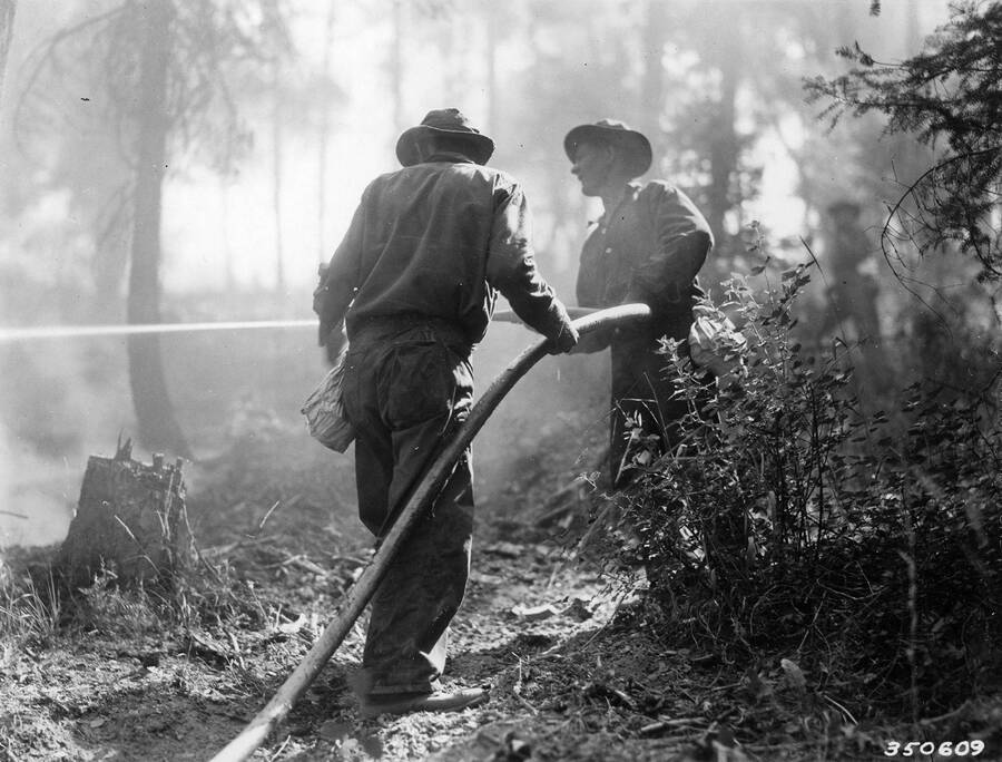 Two CCC men holding a hose in the middle of a smoky forest, presumably fighting a forest fire. Writing beside the photo reads: 'CCC Boys Hosing Down Hotspot'.