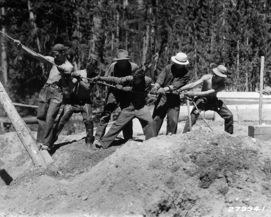 A line of 6 CCC men pull on the same rope. They are standing in a large pile of dirt and seem to be working on some type of work or construction project.