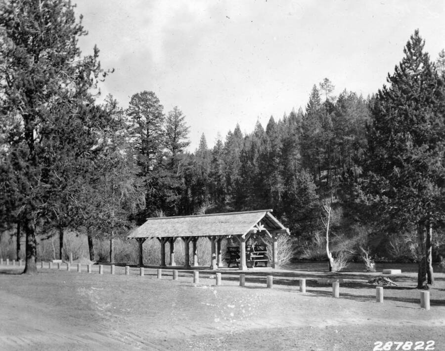 A shelter sits in front of a wooded hill and a line of parking barriers lie in a row in front of it. Underneath the shelter a stack of picnic tables is piled high.