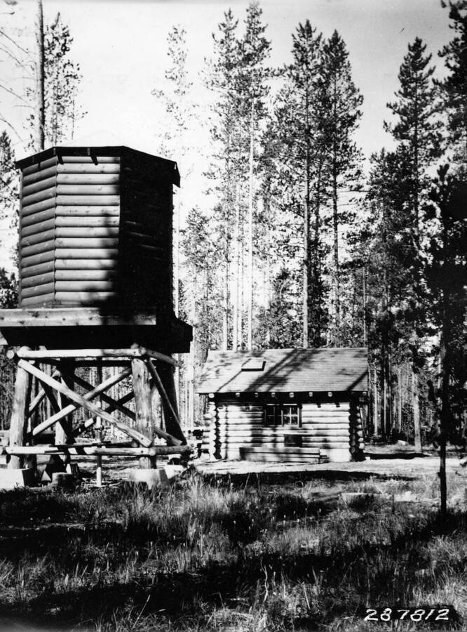 A log cabin sits in a clearing in the woods but is dwarfed by a water tower closer to the foreground.
