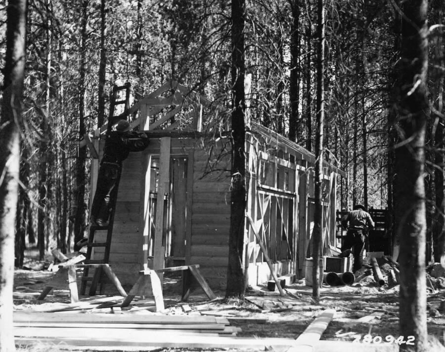 Two CCC men working on the construction of a cabin in the middle of the woods. Two sawhorses are bing used as supports.