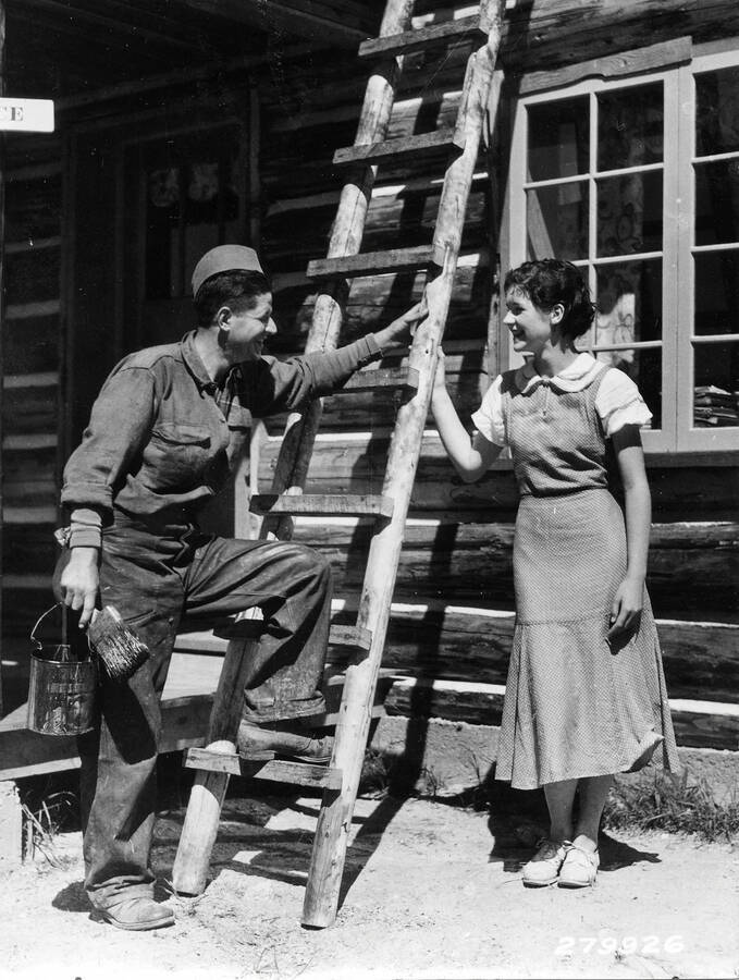A CCC man leans against a ladder holding a paint can and paintbrush. He is smiling at a woman who is also holding onto the ladder. They are both standing in front of a cabin.
