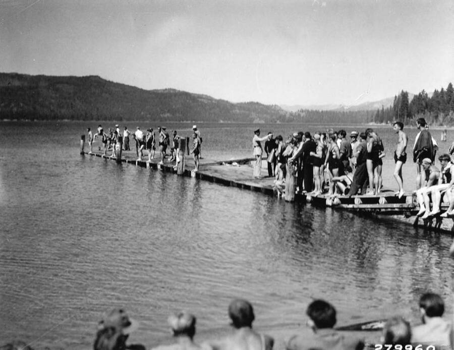 Swimming race contestants posed on the edge of the dock while several other CCC men stand by. Note one man in full military uniform. Spectators can be seen on the shore at the bottom of the photo. Back of photo reads: 'Water Carnival staged by three CCC Camps at McCall, Idaho. Near Idaho National Forest. Taken by K.D. Swan, 1933.'