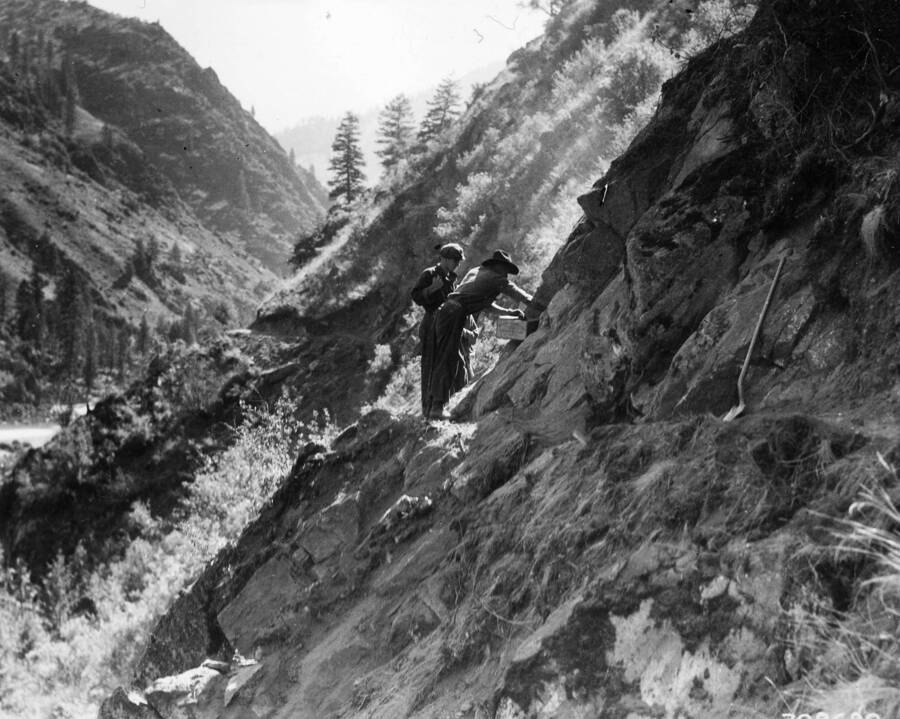 Two CCC men working on the side of a canyon on a road construction project (Presumably the Salmon River road project). On the side of the path a shovel leans against the hill.