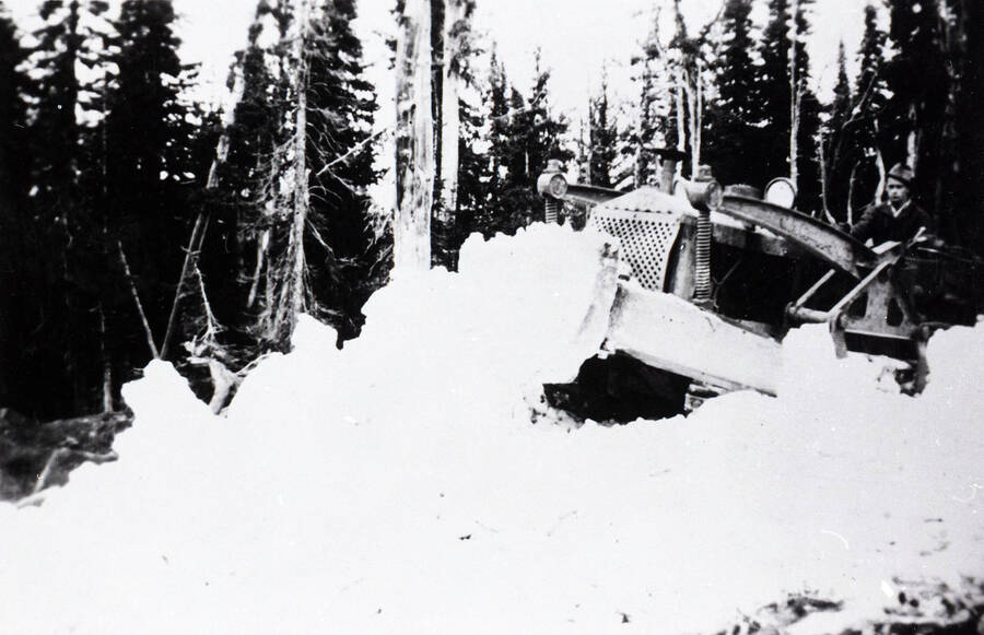 A CCC man drives a bulldozer clearing snow in the forest.
