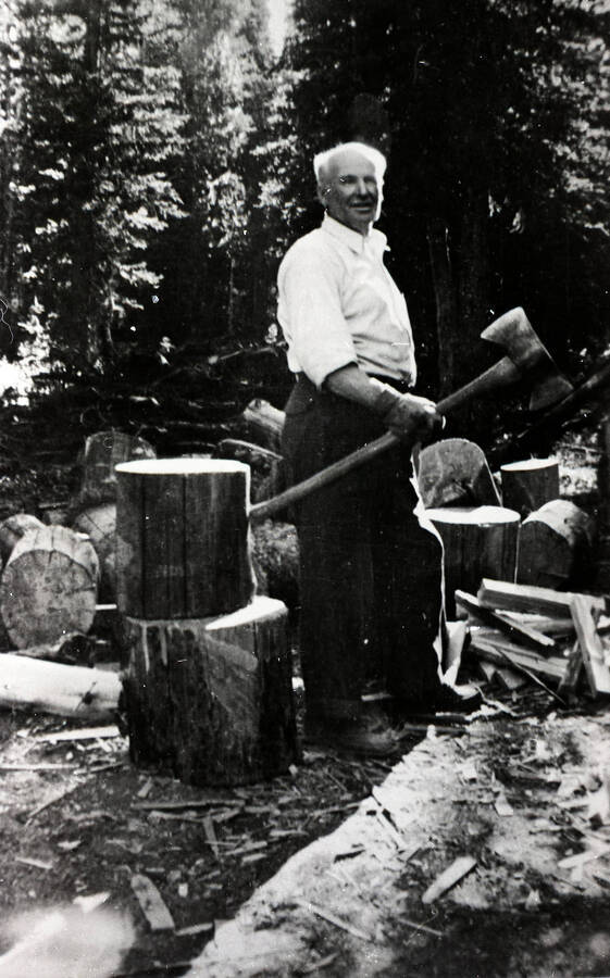 A man stands amongst a pile of stumps and firewood, holding an ax. The picture was taken in the Elk City vicinity.
