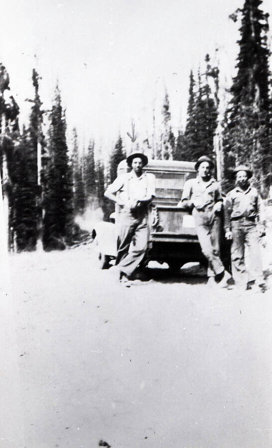Three CCC men pose at the end of the bed of a pickup truck on a dirt road through the forest. There is an arrow that points to the man on the left.