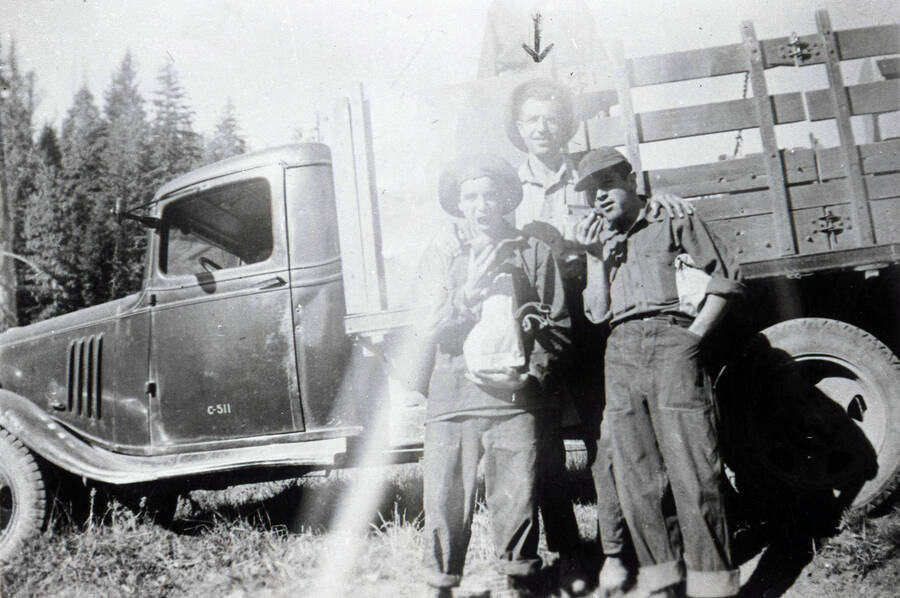 Three CCC enrolles posing in front of truck. Two of the men are pretending to eat food and the last man (standing in the center) has an arrow drawn over his head. The door of the truck has a mark that reads: 'C-511'.