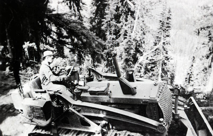 A CCC man drives a bulldozer on the edge of a steep road. The forest falls steeply down the mountainside and another forested hill rises in the background.