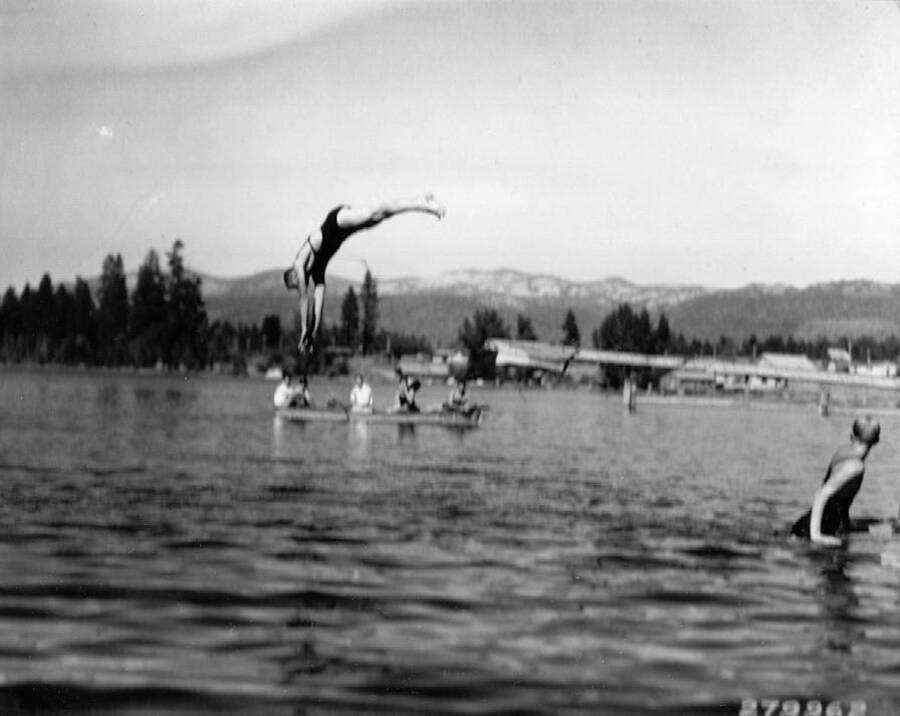 CCC man diving of the end of a diving board into Payette Lake at a CCC water sports carnival diving competition. A group of men can be seen watching in a boat, while another contestant is exiting the water. Back of photo reads: 'Diving Competition CCC water carnival at McCall. Near Idaho National Forest. Taken by K.D. Swan, 1933.'
