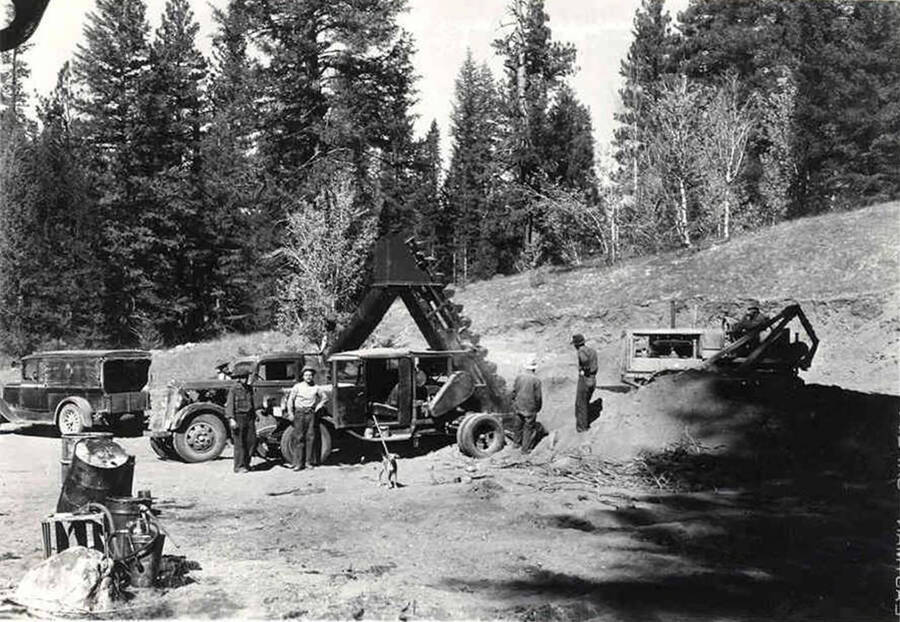 Several CCC men, trucks, a bulldozer, and gravel loader in a quarry with forest in the background.