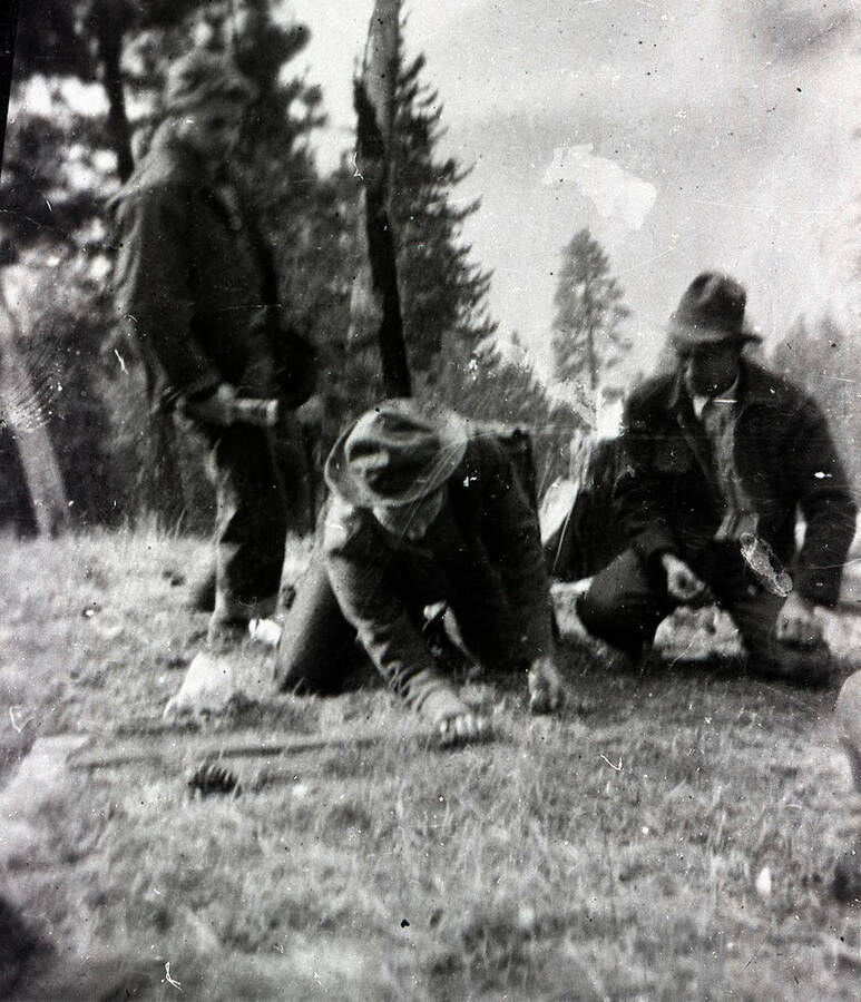 Three CCC men are playing marbles on a grassy hill in the woods. One of the men is measuring the closeness in a marble game with an axe handle.