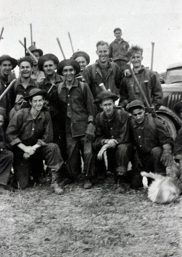A group of CCC enrollees in work uniforms posing for a photo, with shovels and dog.