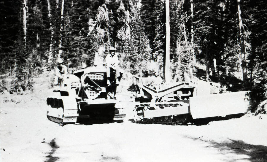 Three CCC men seated on bulldozers on a road in the middle of the forest.