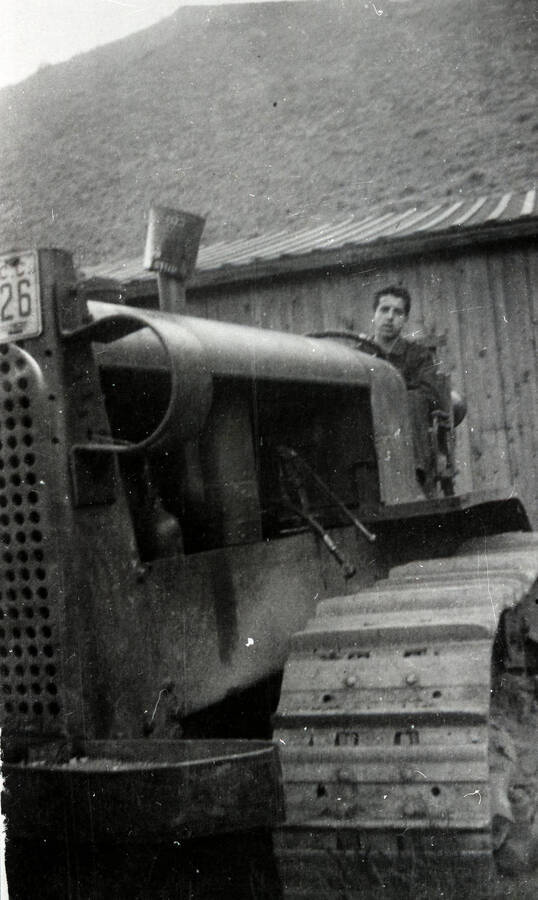 A CCC man sits in the drivers seat of a bulldozer in front of a building and hill.