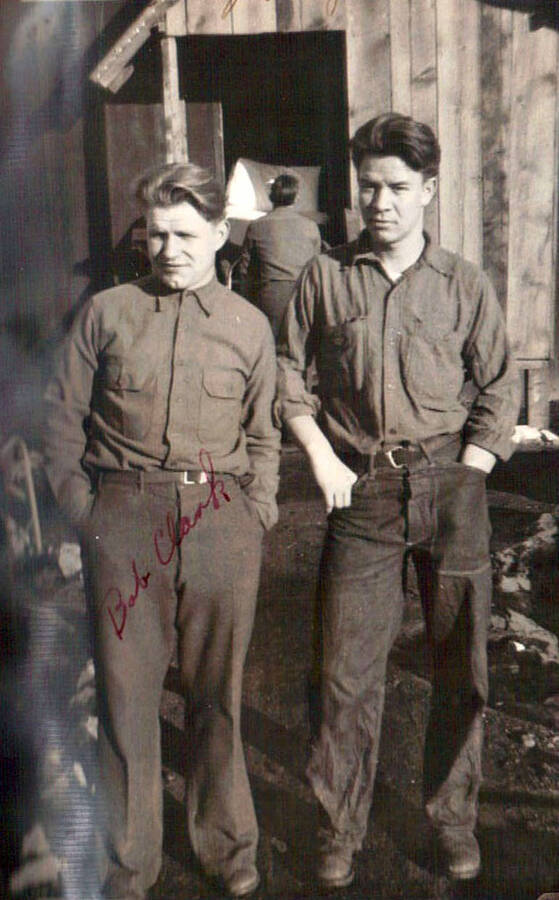 Two CCC men pose in front of a barrack at the CCC camp in Moscow, Idaho. Behind them another CCC man is carrying a bed through the doorway of the barrack. Writing on the photo reads: 'Bob Clark' over the man on the left. Writing above the photo reads: '2 sleepy people (night guards)'.
