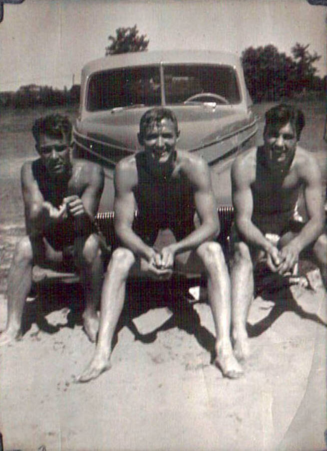 Three CCC Men sitting on a bumper of a car in their swim trunks. Writing that labels an arrow under the photo pointing to the man on the left reads: 'John'.