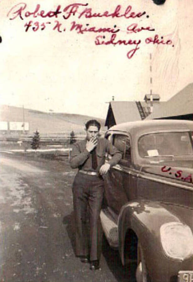 A CCC man leans on a car and appears to be smoking a cigarette. Writing on the photo reads: 'Robert F Buekler 435 N. Miami Ave. Sidney Ohio. U.S.A.'