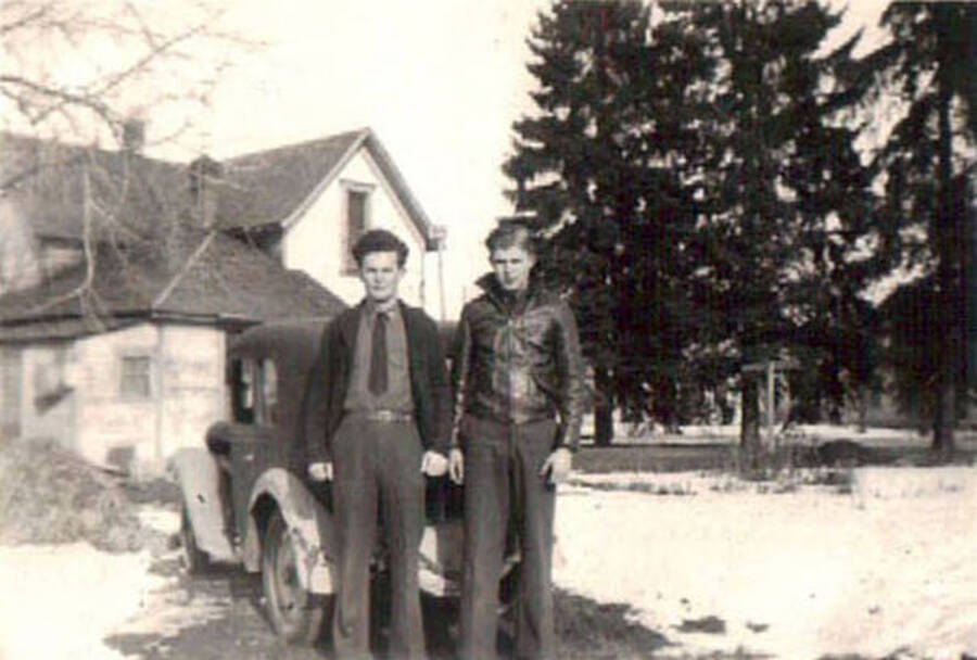 Two CCC men standing in front of a automobile and a house, surrounded by a snow-covered lawn. There are also a few trees in the yard.