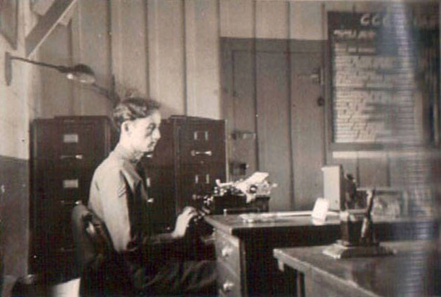 A CCC man sits at a desk with a typewriter. Behind him are filing cabinets, a locked safe, and a chalkboard filled with writing. In the foreground is another desk with what appears to be a pencil sharpener.