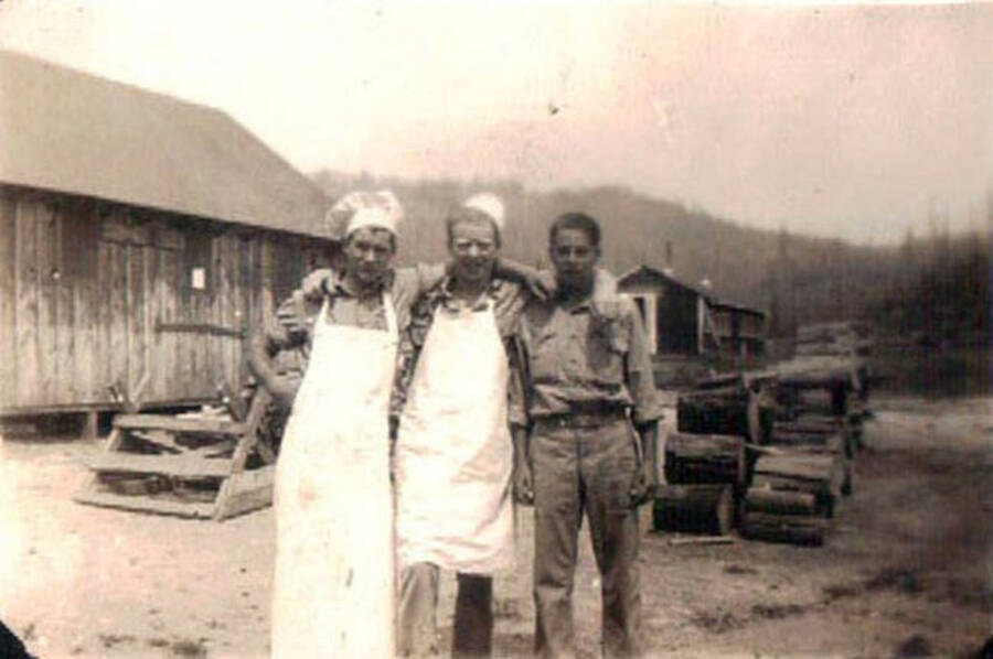 Three CCC men posing in the CCC camp in Moscow, Idaho. The two men on the left are wearing chefs hat and aprons. The men are standing in front of a pile of firewood and a building.