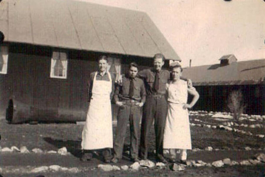 Four CCC men posing on a path in front of a couple buildings in a CCC camp. The two men on the ends are wearing cook's aprons. Writing under the photo reads: 'camp'.