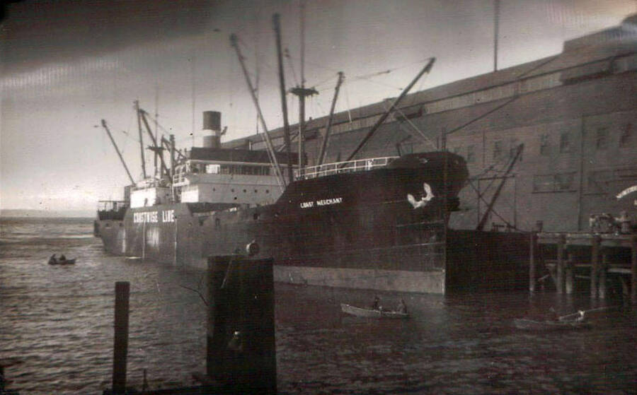An ocean freighter at the dock in Puget Sound. Three small rowboats surround the freighter. The writing on the side of the ship reads: '[Mustise Lite]' and 'L[ea]st Merchant'. Writing under the photo reads: 'Ocean Freighter in Puget Sound. Wash. 1938'.