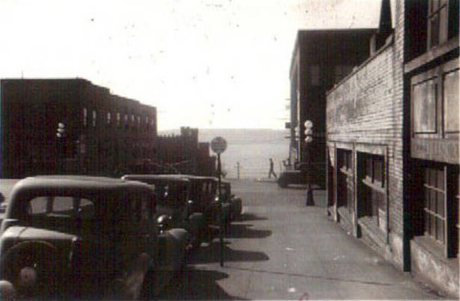 View down a town street with hills in the background. Several parked cars line the road and a pedestrian crosses the street in the distance. Writing under the photo reads: 'Seattle Wash.'