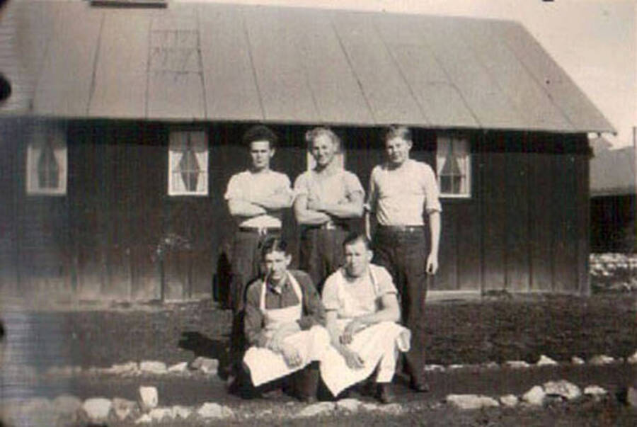 Five CCC men posing on a path in front of a building in a CCC camp in Moscow, Idaho. The two men kneeling on the path are wearing aprons. Writing under the photo reads: 'camp'.