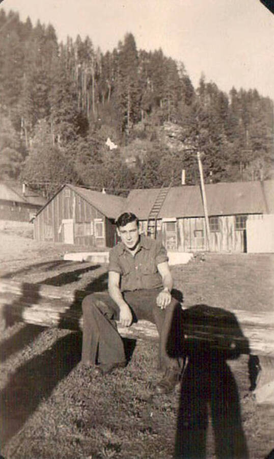 Photo of a CCC man sitting on a log in front of several CCC buildings at the Moscow, Idaho camp. Behind the camp is a forested hill.