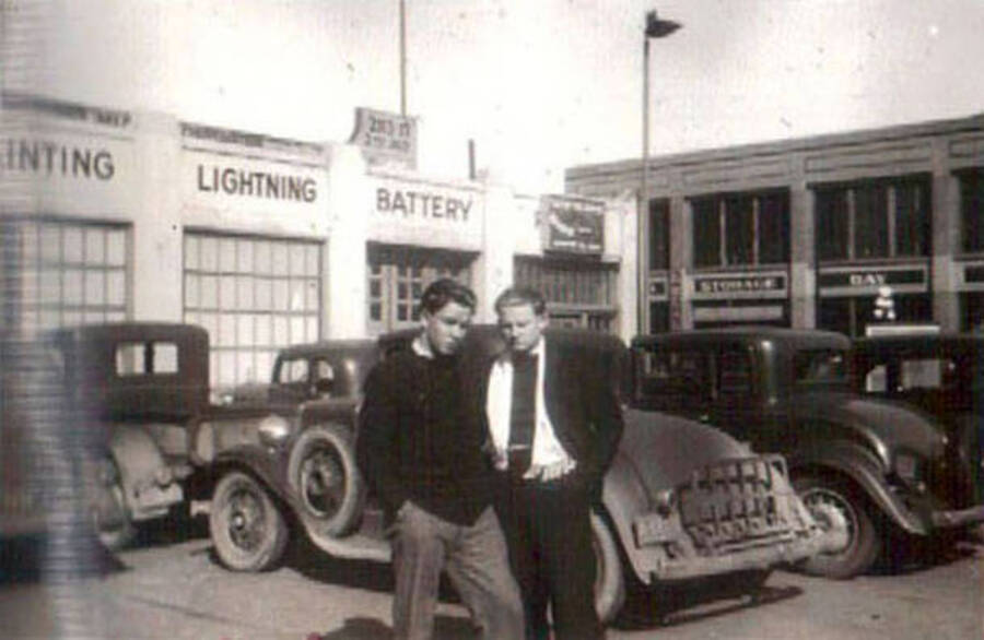 Two men standing together for a photo in a parking lot full of cars in front of a store front. Writing on the building behind the men reads: '[]inting' 'Lightning' 'Battery' 'Storage' 'Bay'. Writing under the photo reads: 'Duck + Brandy'.