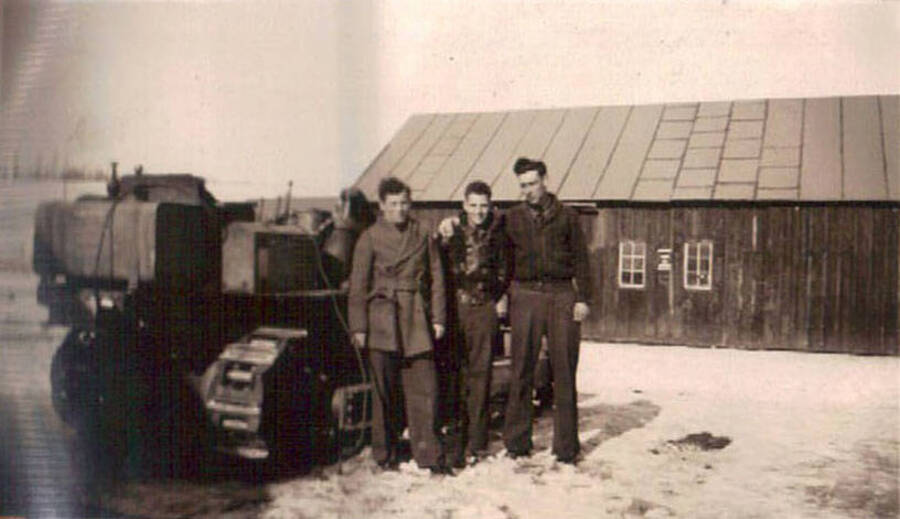 Three CCC men standing in a snowy yard in front of a CCC building. Next to the men is a large bulldozer or snow plow.