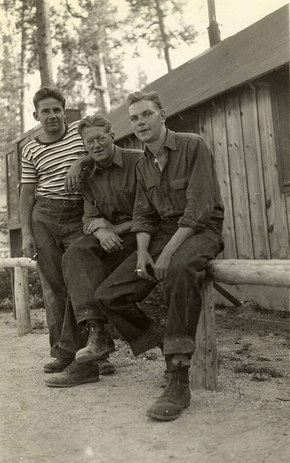 Three CCC men sitting on a log railing in front of a low building in a CCC Camp. Writing below the photo reads: 'Mike and Frank and Me'.