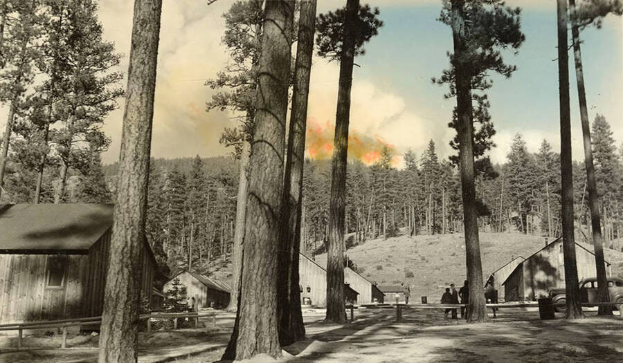 View of a CCC Camp with a fire burning the hills in the background. Writing below the photo reads: 'Fire seen from camp'.