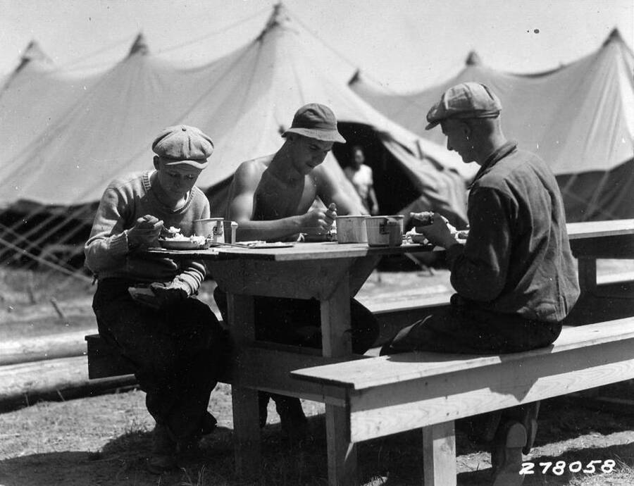 Three CCC men eating lunch at Kaniksu CCC Camp, F-31, Kalispell Bay. Back of photo reads 'Boys at lunch Kaniksu Camp F-31, Kalispell Bay Taken by K.D. Swan.'