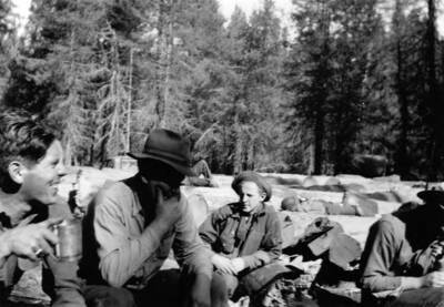 Four CCC men eating, others lie on a field of logs in the background. Writing underneath the photo reads: '1850.0003 CCC (Co. 1997?) Sandy Print'.
