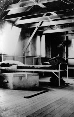 Interior of barrack #5 showing a cot and wooden trunk beneath a window. Writing beneath the photo reads: '1850.0028. Interior of Barracks #5, S-223, McCall, 1935'.